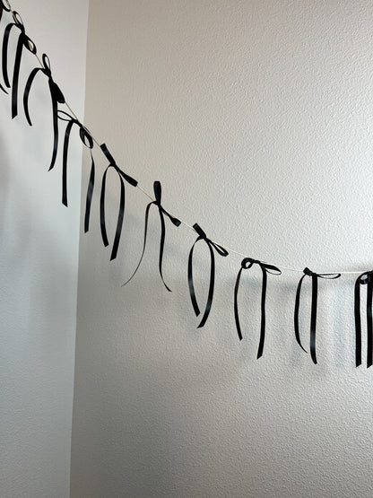 Black ribbon garland with hanging satin bows strung along a cord, displayed against a plain off-white wall.