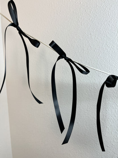 Black satin ribbon garland with tied bows hanging from a thin string against an off-white wall.